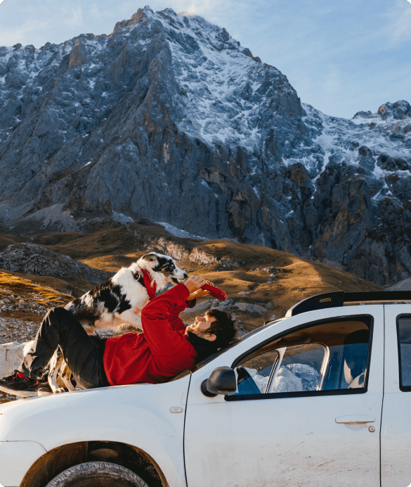 Persoon met hond op een auto met de bergen op de achtergrond.